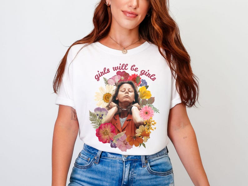 May include: White t-shirt with the text "girls will be girls" in a cursive font. The shirt features a graphic of a person surrounded by colorful flowers. The person is wearing an orange jacket.