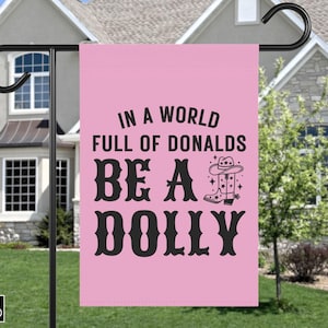 May include: A rectangular pink garden flag featuring black text that reads IN A WORLD FULL OF DONALDS BE A DOLLY. The design includes a black graphic of a cowboy boot with a star pattern and a cowboy hat positioned next to the word DOLLY.