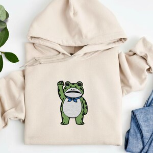 May include: A beige hoodie featuring an embroidered cartoon frog with a blue bow tie. The frog is green with black spots and is raising its right arm. The hoodie has a drawstring hood and long sleeves. The background includes a pair of blue jeans.