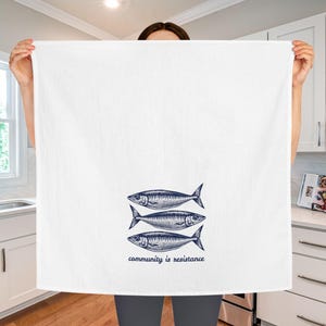May include: White kitchen towel with a navy blue illustration of three fish. The text "community is resistance" is printed below the fish. The towel is held up in front of a kitchen.