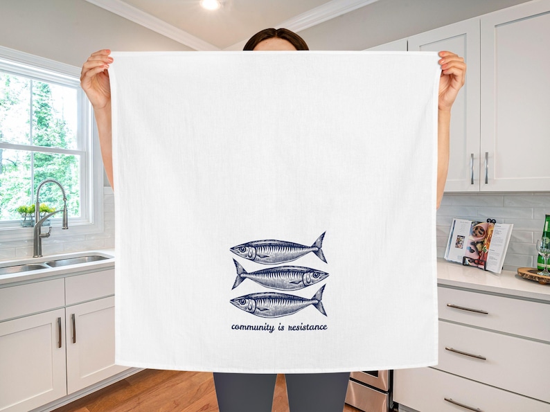 May include: White kitchen towel with a navy blue illustration of three fish. The text "community is resistance" is printed below the fish. The towel is held up in front of a kitchen.