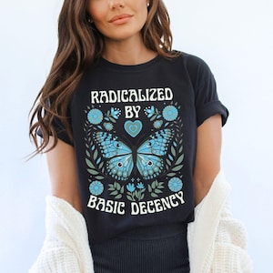 以下が含まれることがあります： 「RADICALIZED BY BASIC DECENCY」の白文字がプリントされた黒いTシャツ。青い蝶のデザインと花のアクセントが特徴です。クルーネックで袖はロールアップされています。