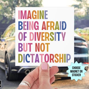 Op de afbeelding: Een witte vierkante magneet of sticker met de tekst "IMAGINE BEING AFRAID OF DIVERSITY BUT NOT DICTATORSHIP" in kleurrijke letters. Een hand houdt het object vast, met een auto op de achtergrond.