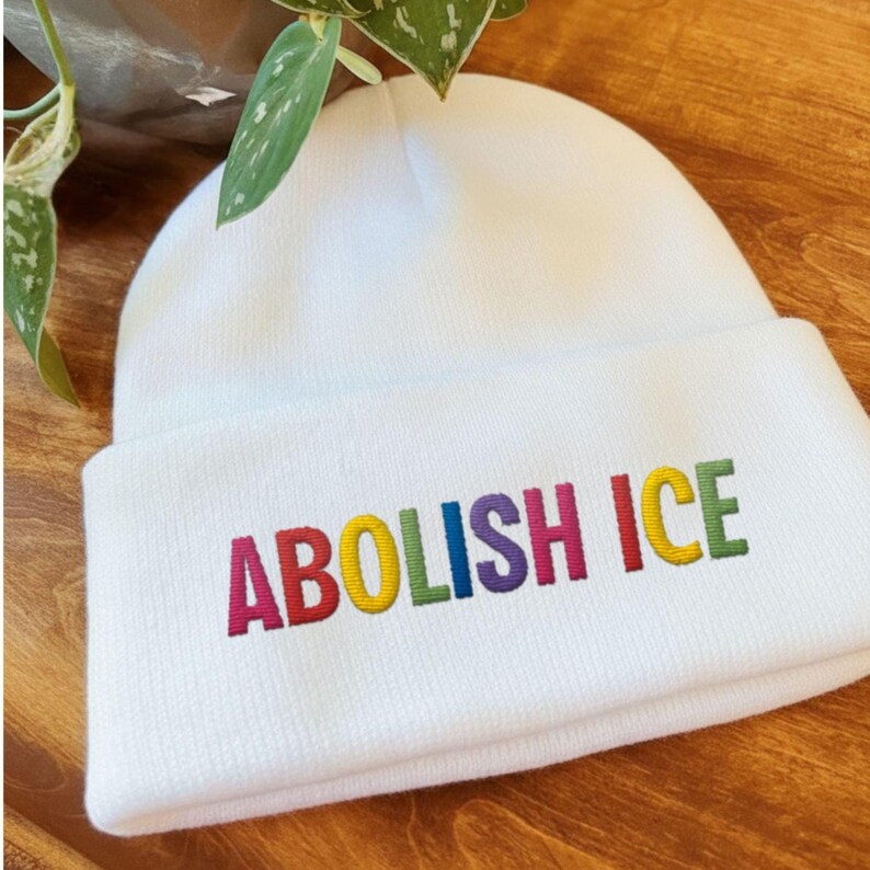 May include: A white beanie with the embroidered text "ABOLISH ICE" in rainbow colors. The hat is made of a soft, knitted material and has a folded brim. The beanie is a casual accessory.