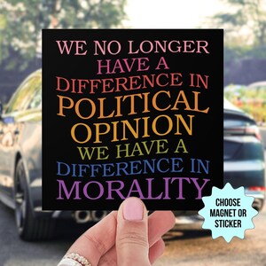 May include: Black square magnet or sticker with the text "WE NO LONGER HAVE A DIFFERENCE IN POLITICAL OPINION WE HAVE A DIFFERENCE IN MORALITY" in colorful letters. A hand holds the magnet in front of a car.