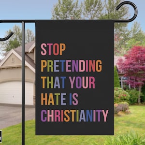 May include: A black garden flag with the text "STOP PRETENDING THAT YOUR HATE IS CHRISTIANITY" in colorful letters. The flag is hanging on a black metal pole in a residential yard.