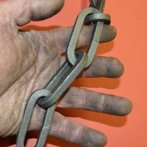 May include: A close-up of a hand holding a heavy-duty metal chain. The chain is made of thick, interlocking links and appears to be used for industrial or construction purposes.
