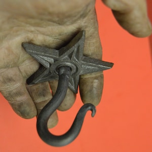 May include: A black metal star-shaped hook with a curved hook at the bottom. The hook is held in a hand.