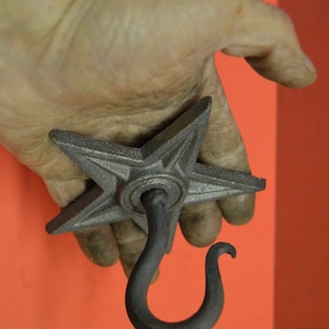 May include: A black metal star-shaped hook with a curved hook at the bottom. The hook is held in a hand.