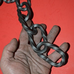 May include: A section of a dark gray metal chain with twisted links. The chain is held in a hand against a red background. The links are thick and have a textured appearance.