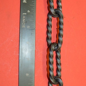 May include: A close-up of a black metal chain with a twisted design. The chain is measured against a ruler, showing it is approximately 12 inches long.