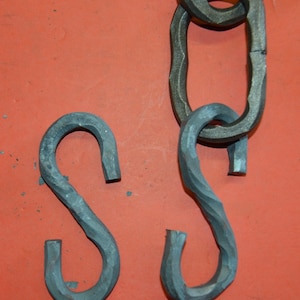 以下が含まれることがあります： ねじれた金属製のS字型のフック2つと、金属製のリンクでできたチェーンの一部。フックとチェーンは赤い表面にあります。