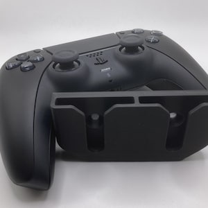 Peut inclure: Une manette de jeu PlayStation 5 DualSense noire avec un support de charge noir. La manette a une poignée texturée et un pavé tactile au centre. Le support a deux emplacements pour maintenir la manette.
