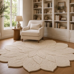 Op de afbeelding: Een crèmekleurig, bloemvormig vloerkleed met een gedetailleerd bloemblaadjesontwerp. Het vloerkleed ligt op een houten vloer in een woonkamer, met een witte fauteuil, een bijzettafel en een boekenkast op de achtergrond.