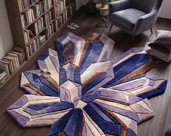 May include: A large, irregularly shaped area rug featuring a geometric pattern of faceted crystal shapes in shades of deep blue, light purple, cream, and brown. The rug has a thick, textured pile and is laid out on a dark wood floor.