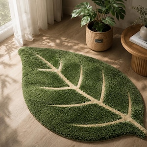 May include: A green leaf-shaped rug with a light beige vein design. The rug is made of a soft, plush material and is placed on a light brown wooden floor. The rug is a decorative item for a living room or bedroom.