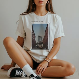 Könnte beinhalten: Weißes T-Shirt mit einem rechteckigen Bild einer Brücke zwischen zwei Gebäuden. Der Text "Ordinary stuff on Film" befindet sich über dem Bild. Die Person trägt schwarz-weiße Turnschuhe und weiße Socken.