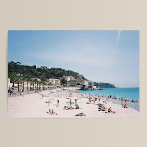 May include: A wide-angle view of a sunny beach with light-coloured sand, numerous people relaxing on the shore, and turquoise water. A promenade with palm trees and buildings lines the coast, backed by a green hill under a clear blue sky.