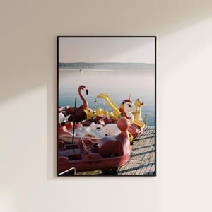 Könnte beinhalten: Ein gerahmter Kunstdruck, der einen Holzsteg mit mehreren bunten Tretbooten in Form eines rosafarbenen Flamingos, eines weißen Einhorns und einer gelben Ente zeigt, die auf ruhigem Wasser vor einem fernen Ufer im klaren Licht treiben.