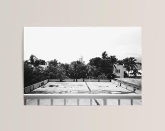Puede incluir: Fotografía en blanco y negro de una azotea con una barandilla en primer plano. Se ven palmeras y otro follaje detrás de la azotea, con edificios y un cielo nublado al fondo.