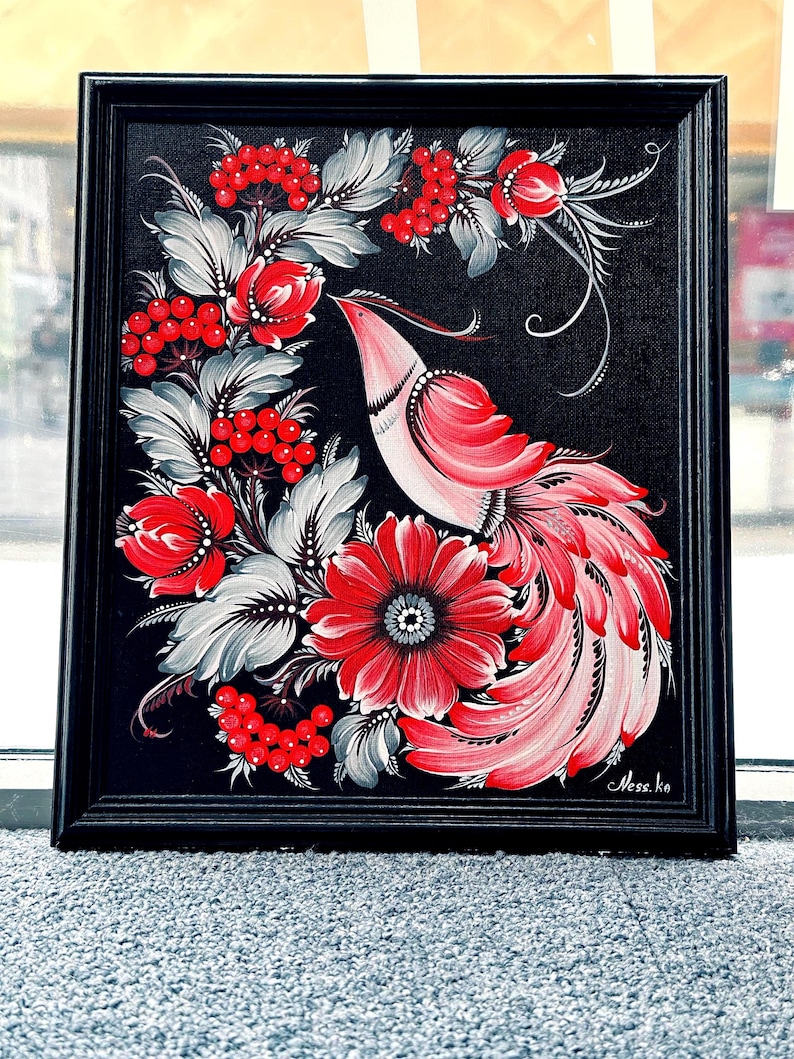 Puede incluir: Pintura enmarcada que presenta un ave estilizada con plumaje rojo y rosa, rodeada de bayas rojas y flores con hojas grises y blancas, sobre un fondo negro. La obra de arte est&aacute; en un marco negro.