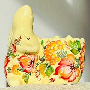 May include: A yellow ceramic bunny-shaped planter, adorned with hand-painted floral designs in red, orange, and green. The planter features a curved back and a scalloped edge, perfect for displaying plants.