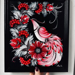 Puede incluir: Pintura enmarcada que representa un p&aacute;jaro estilizado con plumaje rojo y blanco, rodeado de flores rojas, bayas y hojas grises y blancas, sobre un fondo negro. La obra de arte est&aacute; sostenida por una persona.