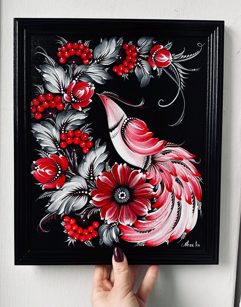 Puede incluir: Pintura enmarcada que representa un p&aacute;jaro estilizado con plumaje rojo y blanco, rodeado de flores rojas, bayas y hojas grises y blancas, sobre un fondo negro. La obra de arte est&aacute; sostenida por una persona.