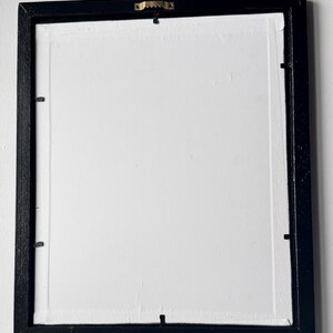 Puede incluir: Parte trasera de una obra de arte enmarcada. El marco negro y rectangular rodea un lienzo blanco. Un colgador de dientes de sierra dorado est&aacute; fijado en el centro superior. Peque&ntilde;os clips met&aacute;licos aseguran el lienzo al marco.