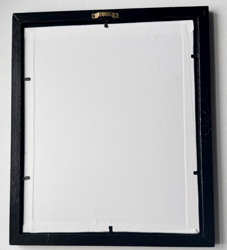 Puede incluir: Parte trasera de una obra de arte enmarcada. El marco negro y rectangular rodea un lienzo blanco. Un colgador de dientes de sierra dorado est&aacute; fijado en el centro superior. Peque&ntilde;os clips met&aacute;licos aseguran el lienzo al marco.