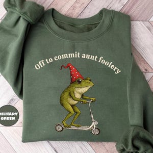 May include: Military green sweatshirt with the phrase "Off to commit aunt foolery" and a graphic of a frog riding a scooter and wearing a party hat. The sweatshirt has long sleeves.