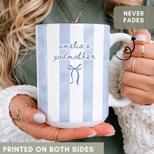 May include: White ceramic mug with blue vertical stripes and the text "amelia's godmother" in a script font. A blue bow graphic is below the text. The mug is printed on both sides and is held by a person.
