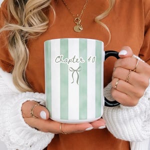 May include: A white ceramic mug with vertical mint green stripes and a black handle. The mug features the text "Chapter 40" with a bow graphic. The mug is held by a person wearing a white sweater.