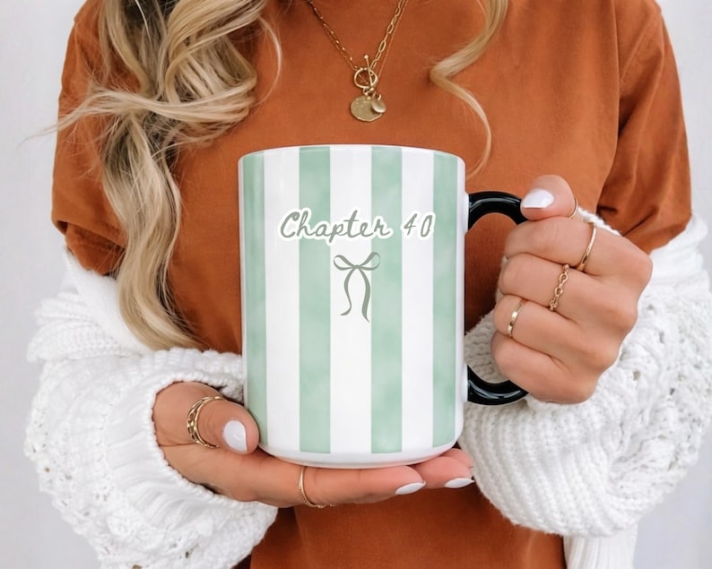 May include: A white ceramic mug with vertical mint green stripes and a black handle. The mug features the text "Chapter 40" with a bow graphic. The mug is held by a person wearing a white sweater.