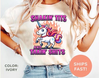 May include: Ivory-colored t-shirt featuring a cartoon unicorn with a blue horn, pink mane, and a star on its flank, set against a fiery background. The shirt displays the text "SHAKIN' TITS AND TAKIN' SHITS" in purple lettering.