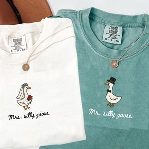 以下が含まれることがあります： クリーム色とティール色の2枚のTシャツ。それぞれに風変わりなガチョウのイラストが描かれています。クリーム色のシャツには、花束を持ったウェディングベール姿のガチョウが描かれ、下に「Mrs. silly goose」と書かれています。ティール色のシャツには、シルクハットと蝶ネクタイをつけたガチョウが描かれ、「Mr. silly goose」と書かれています。