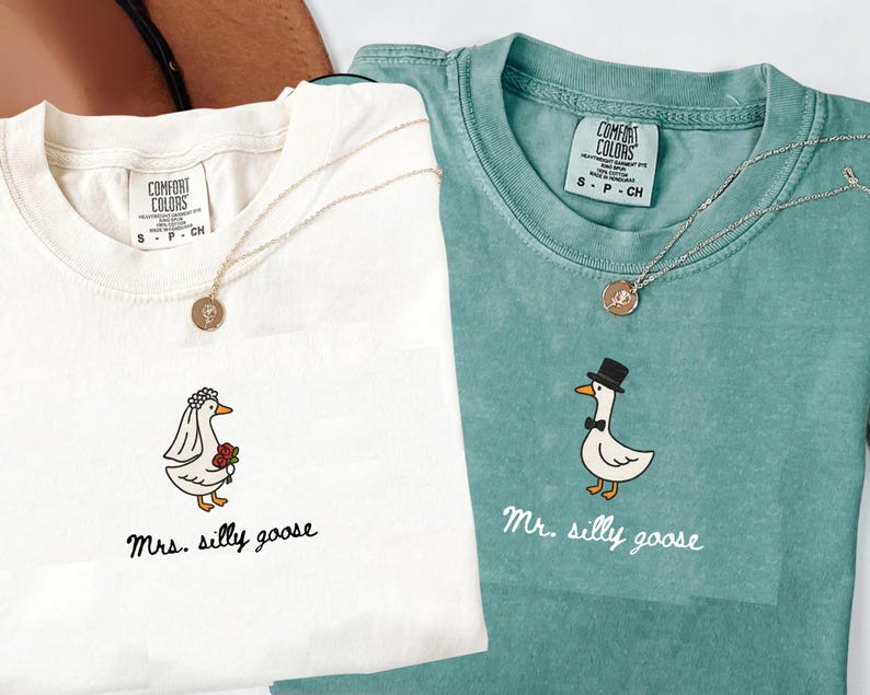以下が含まれることがあります： クリーム色とティール色の2枚のTシャツ。それぞれに風変わりなガチョウのイラストが描かれています。クリーム色のシャツには、花束を持ったウェディングベール姿のガチョウが描かれ、下に「Mrs. silly goose」と書かれています。ティール色のシャツには、シルクハットと蝶ネクタイをつけたガチョウが描かれ、「Mr. silly goose」と書かれています。