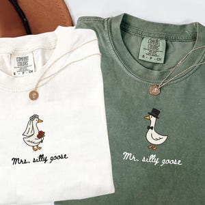 以下が含まれることがあります： クリーム色と緑色の2枚のComfort Colors Tシャツ。それぞれに風変わりなガチョウのグラフィックが描かれています。クリーム色のシャツには「Mrs. silly goose」、緑色のシャツには「Mr. silly goose」と書かれています。どちらのシャツにも金のネックレスが付いています。