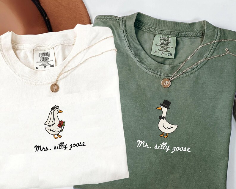 以下が含まれることがあります： クリーム色と緑色の2枚のComfort Colors Tシャツ。それぞれに風変わりなガチョウのグラフィックが描かれています。クリーム色のシャツには「Mrs. silly goose」、緑色のシャツには「Mr. silly goose」と書かれています。どちらのシャツにも金のネックレスが付いています。