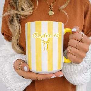 May include: White ceramic mug with yellow stripes and a yellow handle. The mug has the text "Chapter 40" with a bow graphic. The mug is held by a person wearing a brown shirt and a white sweater.
