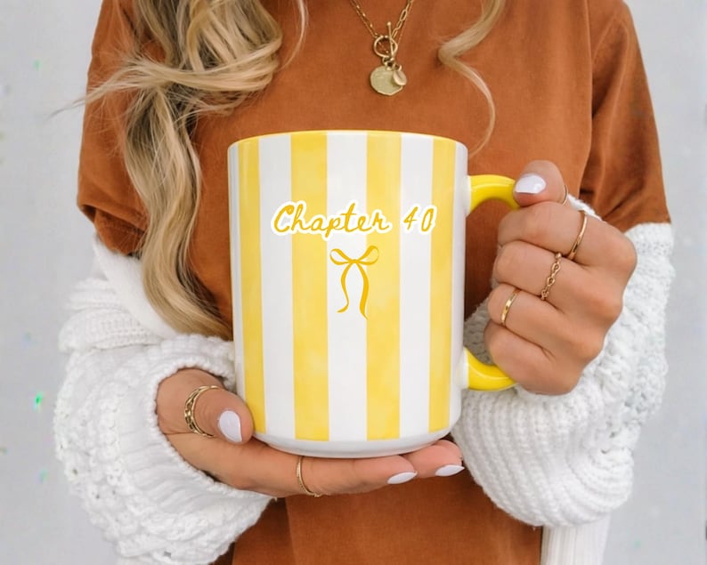 May include: White ceramic mug with yellow stripes and a yellow handle. The mug has the text "Chapter 40" with a bow graphic. The mug is held by a person wearing a brown shirt and a white sweater.