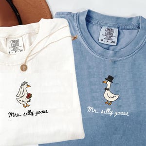 以下が含まれることがあります： 白と青の2枚のComfort Colors Tシャツ。お揃いの刺繍ガチョウのデザイン。白いシャツには「Mrs. silly goose」、青いシャツには「Mr. silly goose」と書かれています。金のネックレスが白いシャツの上にあります。