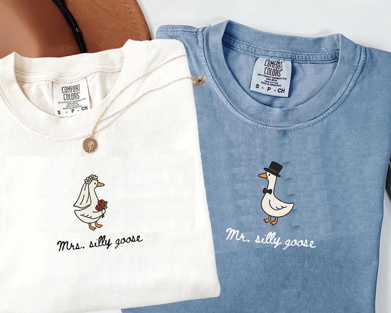 以下が含まれることがあります： 白と青の2枚のComfort Colors Tシャツ。お揃いの刺繍ガチョウのデザイン。白いシャツには「Mrs. silly goose」、青いシャツには「Mr. silly goose」と書かれています。金のネックレスが白いシャツの上にあります。