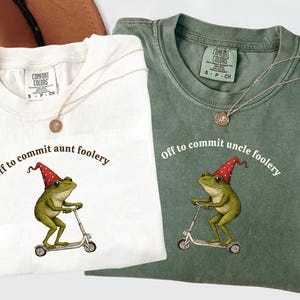 May include: Two t-shirts, one cream and one green, with a frog graphic riding a scooter and wearing a red party hat. The text "Off to commit aunt foolery" and "Off to commit uncle foolery" are printed on the shirts. Each shirt has a gold necklace.