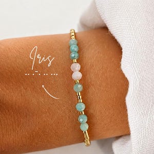 Peut inclure: Un bracelet de perles composé de pierres facettées bleu clair et rose pâle alternées avec de petites perles métalliques dorées. Le mot Iris est écrit en écriture blanche à côté du bracelet avec une ligne pointillée et une flèche.