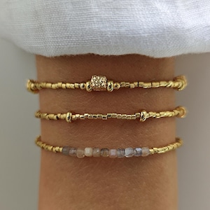 Peut inclure: Trois bracelets dorés superposés sur un poignet. Le bracelet supérieur a un centre carré texturé. Le bracelet du milieu a de petites perles et des accents dorés. Le bracelet inférieur présente de petites pierres carrées de couleur claire.