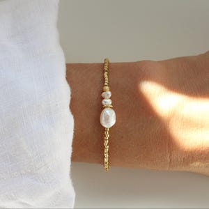 Peut inclure: Un bracelet délicat composé d'une grande perle baroque blanche centrale entourée de deux perles blanches rondes plus petites, agrémenté de petites perles métalliques dorées disposées en motif linéaire le long du bijou.