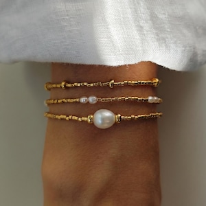 Peut inclure: Un ensemble de trois bracelets délicats composés de petites perles cylindriques dorées. Le bracelet supérieur est entièrement doré, celui du milieu intègre de minuscules perles blanches, et le bracelet inférieur présente une perle blanche centrale.