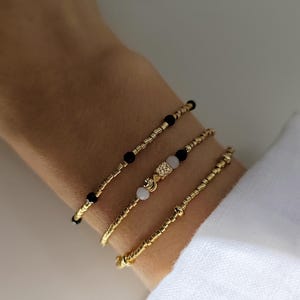 Könnte beinhalten: Ein Set aus drei Perlenarmbändern mit goldfarbenen zylindrischen Perlen. Ein Armband enthält schwarze kugelförmige Perlen, ein weiteres zeigt eine zentrale goldfarbene texturierte Würfelperle zwischen zwei hellrosa Perlen, das dritte ist einfarbig goldfarben.