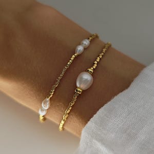 Op de afbeelding: Twee gouden kralenarmbanden met witte parels. Eén armband heeft kleine parels, de andere een grote centrale parel. De armbanden worden om een pols gedragen, waarbij het ontwerp en de textuur van de sieraden worden getoond. Een delicaat accessoire.